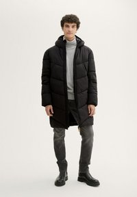 Manteau doudoune noir avec capuche, design matelassé, coupe longue, fermeture éclair devant, texture superposée, porté sur un pull à col roulé gris, associé à un jean noir et des bottes.