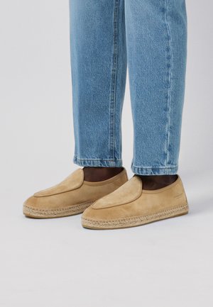 Pies con mocasines sin cordones de ante color beige con suelas trenzadas, combinados con vaqueros azul claro de pierna recta sobre un fondo gris.