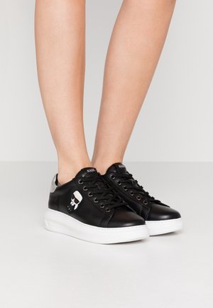 Osoba nosząca czarne skórzane trampki z białymi podeszwami i małym kreskówkowym logo po boku, stojąca na jednolitej jasnej podłodze.