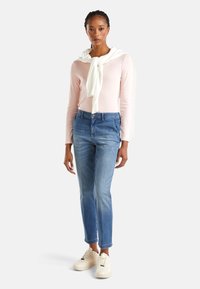 Maglietta a maniche lunghe rosa chiaro con un maglione bianco annodato al collo, jeans blu slim-fit e sneakers beige con dettagli testurizzati.