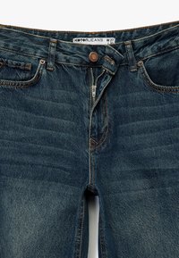 Jeans de mezclilla azul oscuro con cremallera y cierre de botón. Incluye cinco bolsillos y costuras visibles a lo largo de las costuras.