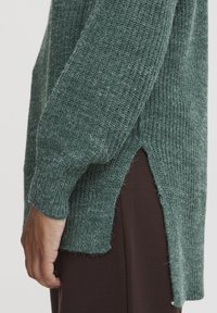 Grüner gestrickter Pullover mit gerippter Textur, seitlichen Schlitzen und eng anliegenden Bündchen, kombiniert mit dunkelbraunen Hosen, die eine lockere, lässige Passform zeigen.