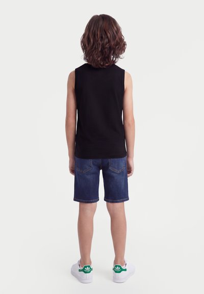 Παιδί με μεσαίου μήκους σγουρά μαλλιά, που φοράει μαύρο αμάνικο πουκάμισο, μπλε τζιν σορτς και λευκά αθλητικά παπούτσια, με την πλάτη προς το φακό σε λευκό φόντο.