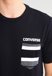 Černé tričko s malou černobílou texturovanou kapsou a bílým logem "CONVERSE" nad kapsou na levé části hrudi.