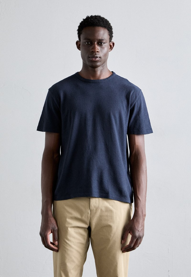 T-shirt en coton bleu marine à manches courtes, col rond et coupe décontractée, porté avec un pantalon beige clair. Texture douce, design minimal.