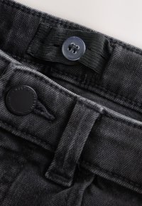 Adjustable black denim jeans waistband with a button and elastic strap for size customization.