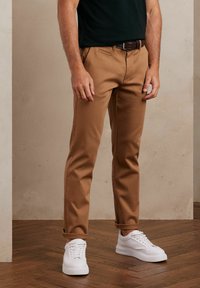 Brązowe spodnie slim-fit wykonane z tkaniny tkanej, z bocznymi kieszeniami i podwiniętymi mankietami, zestawione z białymi trampkami. Gładka tekstura, czyste linie.