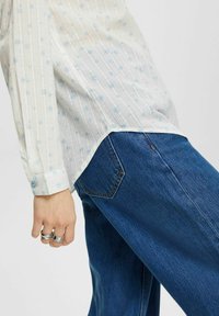 Camisa blanca de manga larga con un patrón floral azul claro y rayas verticales, combinada con Jeans de mezclilla azul. Presenta puños abotonados y un corte relajado.