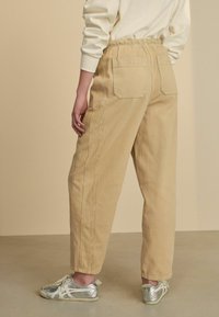 Pantalons en velours côtelé beige avec des rayures verticales, dotés de poches arrière et d'une taille élastique, portés avec des chaussures de sport argentées.