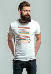 Weißes T-Shirt mit einem farbenfrohen abstrakten Design in orangefarbenen, blauen und grünen Streifen, mit dem Schriftzug "SOUTH BEACH" darunter. Schwarze Jeans.