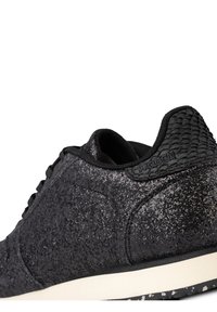 Gros plan d'une basket noire avec un tissu texturé et un contre-renfort de talon à motifs au-dessus d'une semelle blanche et noire tachetée.