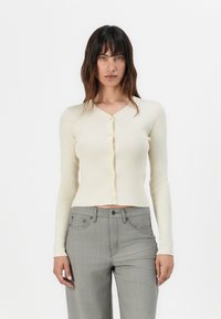 Cardigan a coste crema con maniche lunghe, scollatura a V e chiusura con bottoni. Abbinato a pantaloni grigi a vita alta. Texture morbida e design aderente.