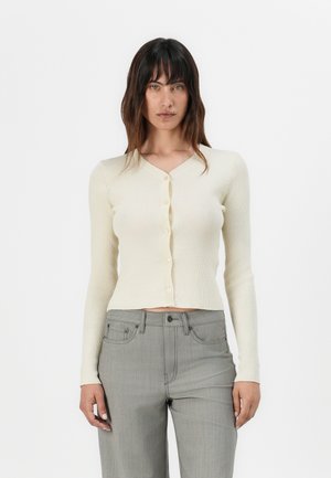 Cardigan a coste crema con maniche lunghe, scollatura a V e chiusura con bottoni. Abbinato a pantaloni grigi a vita alta. Texture morbida e design aderente.