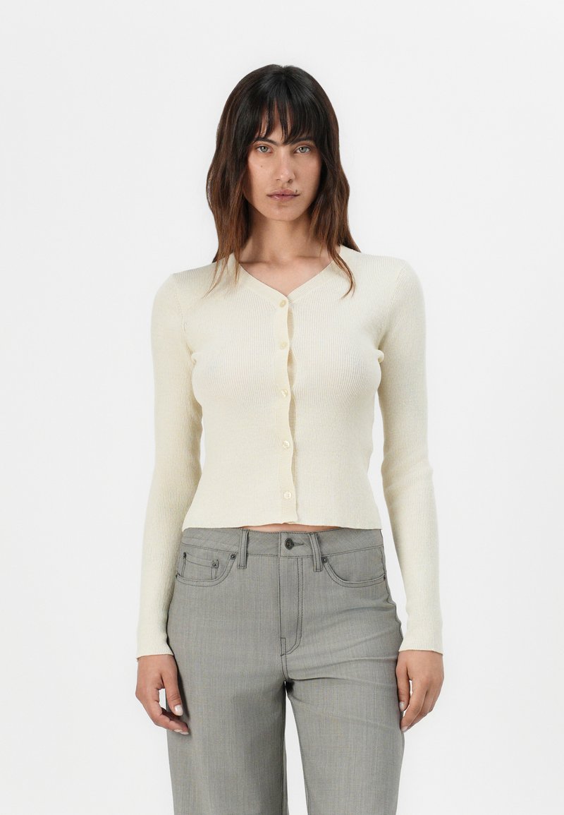 Cardigan a coste crema con maniche lunghe, scollatura a V e chiusura con bottoni. Abbinato a pantaloni grigi a vita alta. Texture morbida e design aderente.