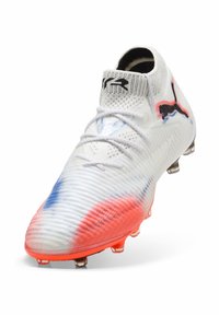Scarpa da calcio bianca con superficie texture, caratterizzata da dettagli blu e rossi, logo Puma nero e tacchetti trasparenti per una migliore trazione.