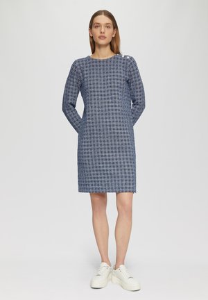 Marineblaues, strukturiertes Kleid mit langen Ärmeln, seitlichen Taschen und rundem Ausschnitt. Verfügt über einen Knopfakzent auf der Schulter. Getragen mit weißen Sneakers.
