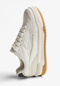 Tenisky s bielou koženou hornou časťou, zaoblenou špičkou, gumovou podrážkou a gumovým vonkajším plášťom. Obsahujú ploché šnúrky a perforované bočné detaily.