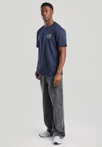 Camiseta de algodón azul marino con mangas cortas y cuello redondo, con un pequeño gráfico blanco en el pecho, combinada con unos pantalones vaqueros de pierna ancha en gris desgastado.