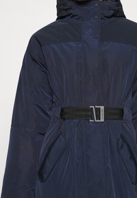 Granatowa parka z kapturem, z czarnym paskiem i dwoma bocznymi kieszeniami. Materiał ma gładkie, błyszczące wykończenie.