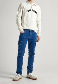 Camisa polo blanca de manga larga con "PEPE JEANS" en negro, combinada con jeans cargo azules y zapatillas beige con cordones blancos y suelas de goma.