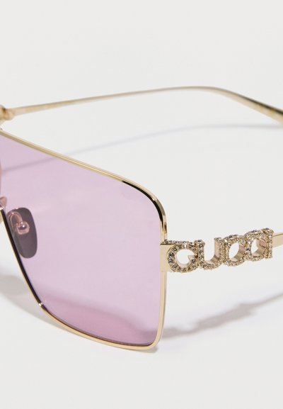 Kultakehyksiset aurinkolasit, joissa on vaaleanpunaiset sävylinssit. Sivussa suuri, korukivillä koristeltu "Gucci"-logo. Tyylikäs, suorakaiteenmuotoinen muotoilu.