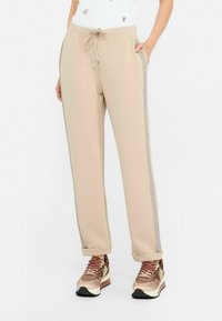 Pantalons de survêtement beiges avec une taille à cordon, dotés de bandes latérales texturées argentées et de poignets élastiques. Associés à des baskets marron et or.