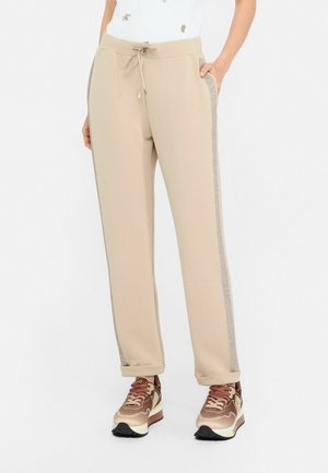 Pantaloni sportivi beige con vita regolabile, dotati di strisce laterali testurizzate in argento e polsini elasticizzati. Abbinati a sneakers marroni e dorate.