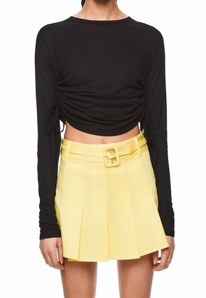 Frau trägt ein langärmliges, schwarzes, bauchfreies Oberteil mit seitlichen Bändern und einen hoch taillierten, gelben, plissierten Rock mit passendem Gürtel.