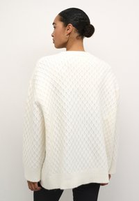 Witte gebreide trui met een ruitpatroon, lange mouwen en een geribde zoom. Zachte textuur en een oversized pasvorm, met minimale details.
