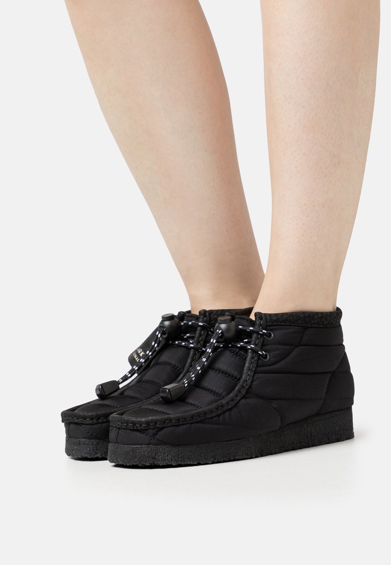 Πόδια που φορούν μαύρες επενδεδυμένες αστραγάλες μπότες με παχιά σόλες και κορδόνια με κούμπωμα τύπου toggle σε λευκό φόντο.