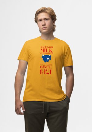 Gul bomulds T-shirt med blå tekst "LAVET MED mælk" og en blå ko grafik, med røde detaljer og året "1921." Korte ærmer.