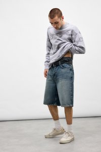 Ung mand i en grå sweater med kædemønster løfter den, iført shorts af denim til knæene, hvide sokker og beige sneakers på et ensfarvet gråt gulv.