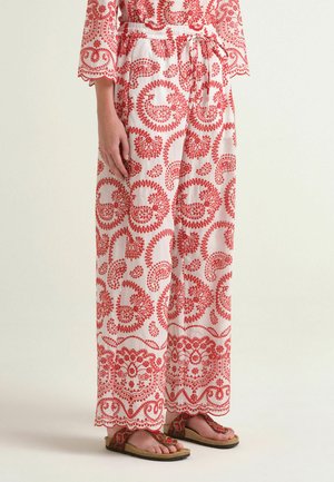 Pantaloni bianchi a gamba larga con motivi paisley rossi e vita con coulisse, indossati con sandali rossi e dorati.