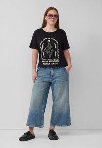 Zwarte katoenen t-shirt met een grafische print, gecombineerd met losse lichtblauwe denim culottes en zwarte instappers. Grote ronde zonnebril gedragen.