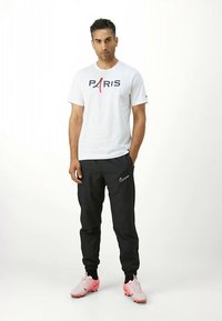 Balti marškinėliai su užrašu „PARIS“, juodos laisvalaikio kelnės su „Nike“ logotipu ir rožiniai sportbačiai, stovintys priešais šviesų foną.