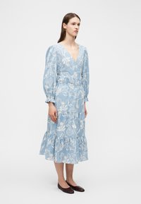 Femme portant une robe midi bleu clair avec une broderie florale blanche, des manches longues bouffantes, un décolleté en V profond, et des ballerines marron foncé, debout immobile.