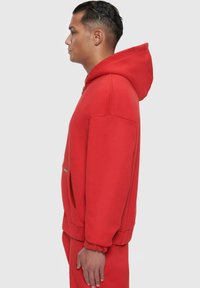 Haut à capuche rouge avec une poche avant, des poignets et un ourlet côtelés. Tissu doux ; coupe oversize. La capuche est attachée et le design est simple et épuré.