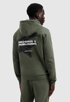 Man in een olijfgroene hoodie en joggingbroek kijkt zijwaarts, op de achterkant van de hoodie staat 'RECORDS & PUBLISHING' geschreven over zwarte verfstrepen.