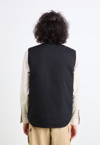 Schwarze ärmellose Weste mit glatter Textur, geradem Design und abgerundetem Saum. Über einem hellen Langarmshirt getragen, von hinten gesehen.