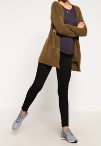 Brązowy sweter z dzianiny z otwartym przodem i kieszeniami, połączony z fioletową bluzką, czarnymi obcisłymi dżinsami i szarymi butami sportowymi.