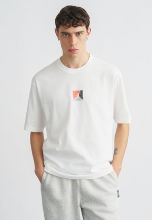 Άσπρο βαμβακερό t-shirt με κοντά μανίκια, που απεικονίζει ένα μικρό, πολύχρωμο αφηρημένο σχέδιο σε κόκκινο, μαύρο και γκρι στο στήθος.