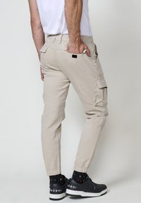 Beige cargo nadrág oldal zsebekkel és szűkített szárakkal, pamut anyagból készült. Fekete sportcipővel és mintás zoknival viselve.
