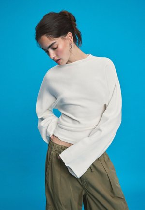 Jeune femme aux cheveux bruns attachés porte un haut blanc à manches longues et un pantalon vert olive sur fond bleu.