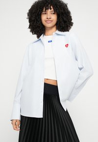 Svetlomodrá pruhovaná košeľa s červeným srdcovým logom na hrudi, doplnená bielym krátkym topom a čiernou plisovanou sukňou. Hladká textúra, voľný strih.