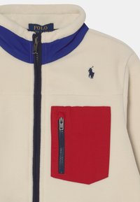 Kremowa polarowa kurtka z wysokim kołnierzem, fioletowymi akcentami i czerwoną kieszenią na zamek błyskawiczny. Posiada małe granatowe logo na klatce piersiowej.