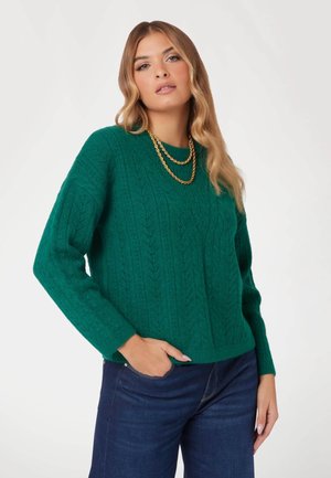 Green knitted sweater with a textured cable design, long sleeves, and a crew neckline, paired with dark blue jeans. Gold chain necklace accent.