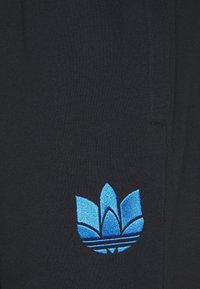 Svarta joggers av tyg med en blå broderad logotyp med tre stiliserade löv och horisontella ränder nära basen. Slät textur.