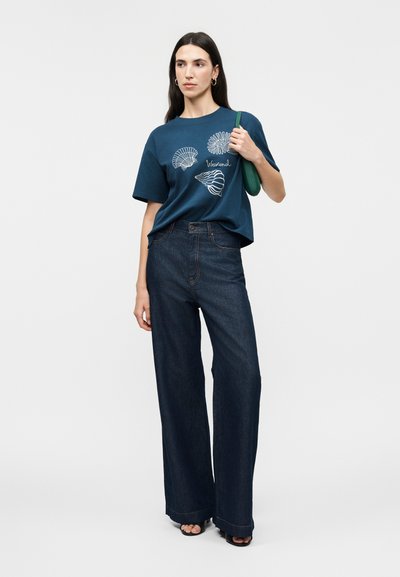 Γυναίκα που φοράει μπλε μπλουζάκι με λευκά σχέδια από κοχύλια και το κείμενο «Weekend», φαρδιά σκούρα τζιν, μαύρα σανδάλια και κρατάει μια μικρή πράσινη τσάντα ώμου.