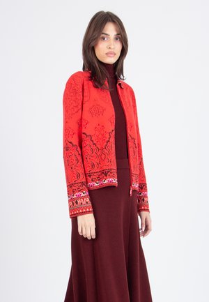 Veste rouge avec des motifs noirs complexes et des accents brodés multicolores aux poignets, associée à un col roulé bordeaux et une jupe.