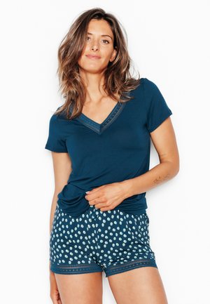 Vrouw in een marineblauw kortermig shirt en marineblauwe korte broek met polkadots en kanten rand, staand tegen een witte achtergrond.